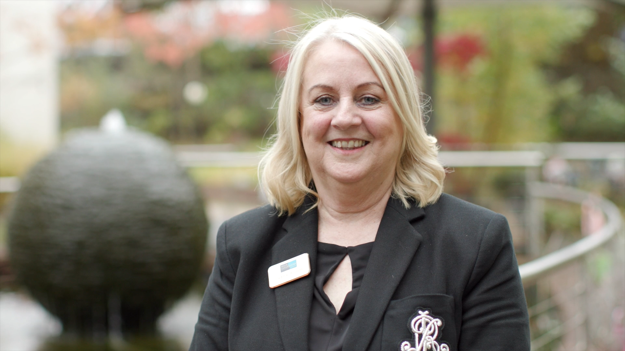 A lady with light hair and a dark suti wearing a nametag in a garden with a water feature