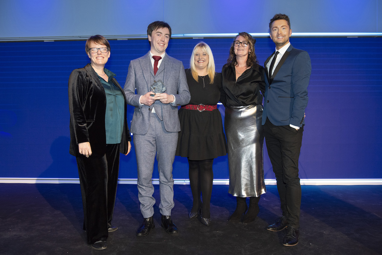 A man in a grey suit holds his glass trophy whilst smiling. Flanked by three women in evening wea and a man in a dark suit