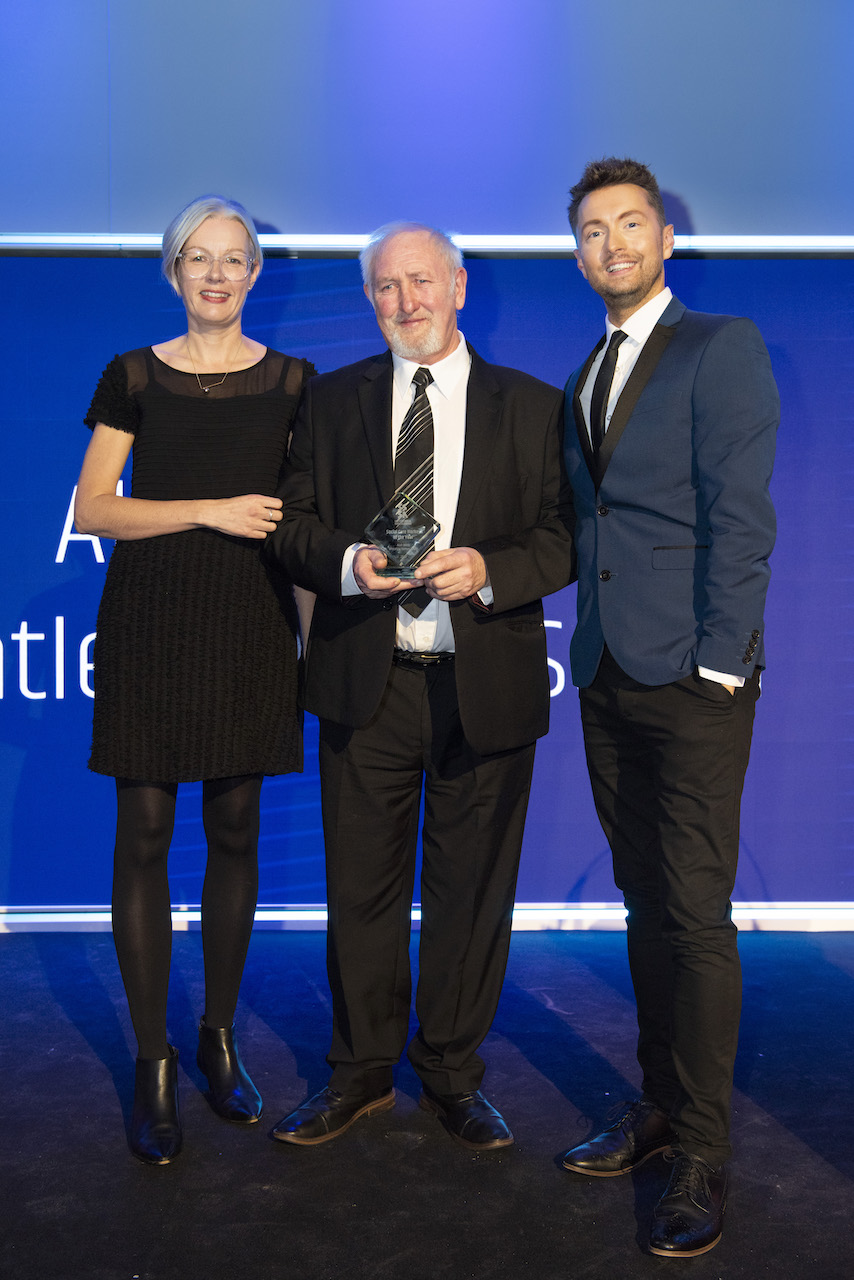 a man in a light suit holds his glass award smiling, between a woman in dark evening wear and a man in a dark suit