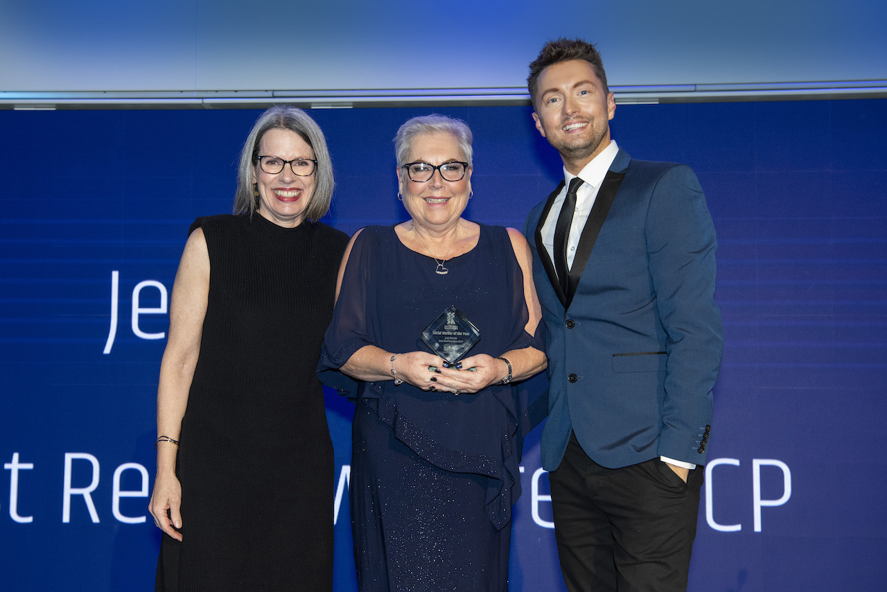 A woman in blue evening wear holds her glass trophy smiling, with a woman in dark evening wear and a man in a blue suit