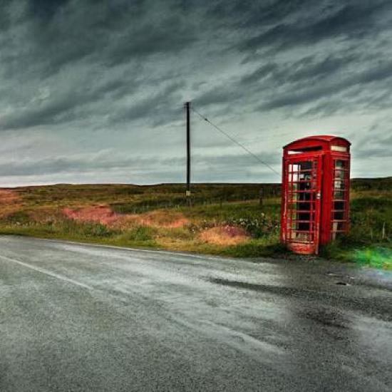 Old red phone kiosk by rural road under looming skies