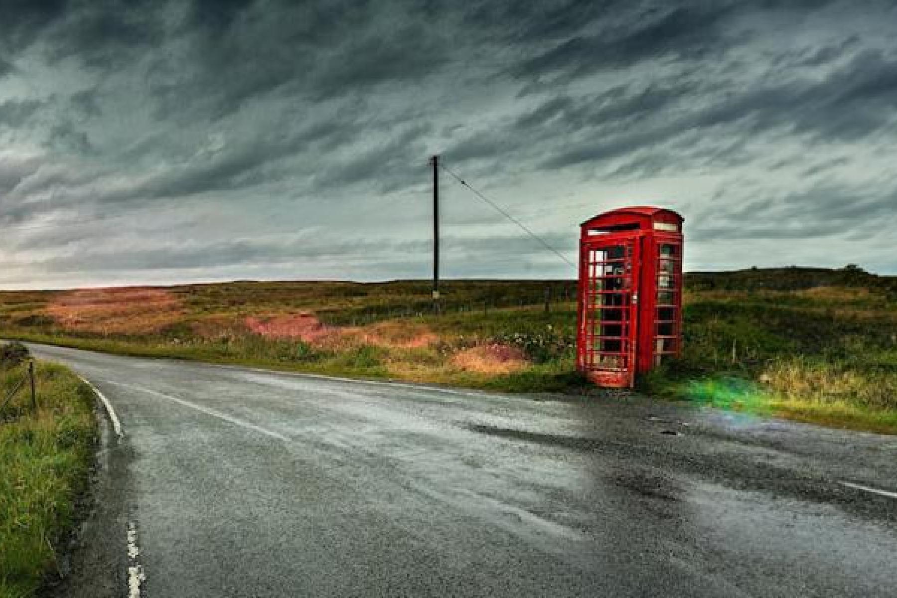 Old red phone kiosk by rural road under looming skies