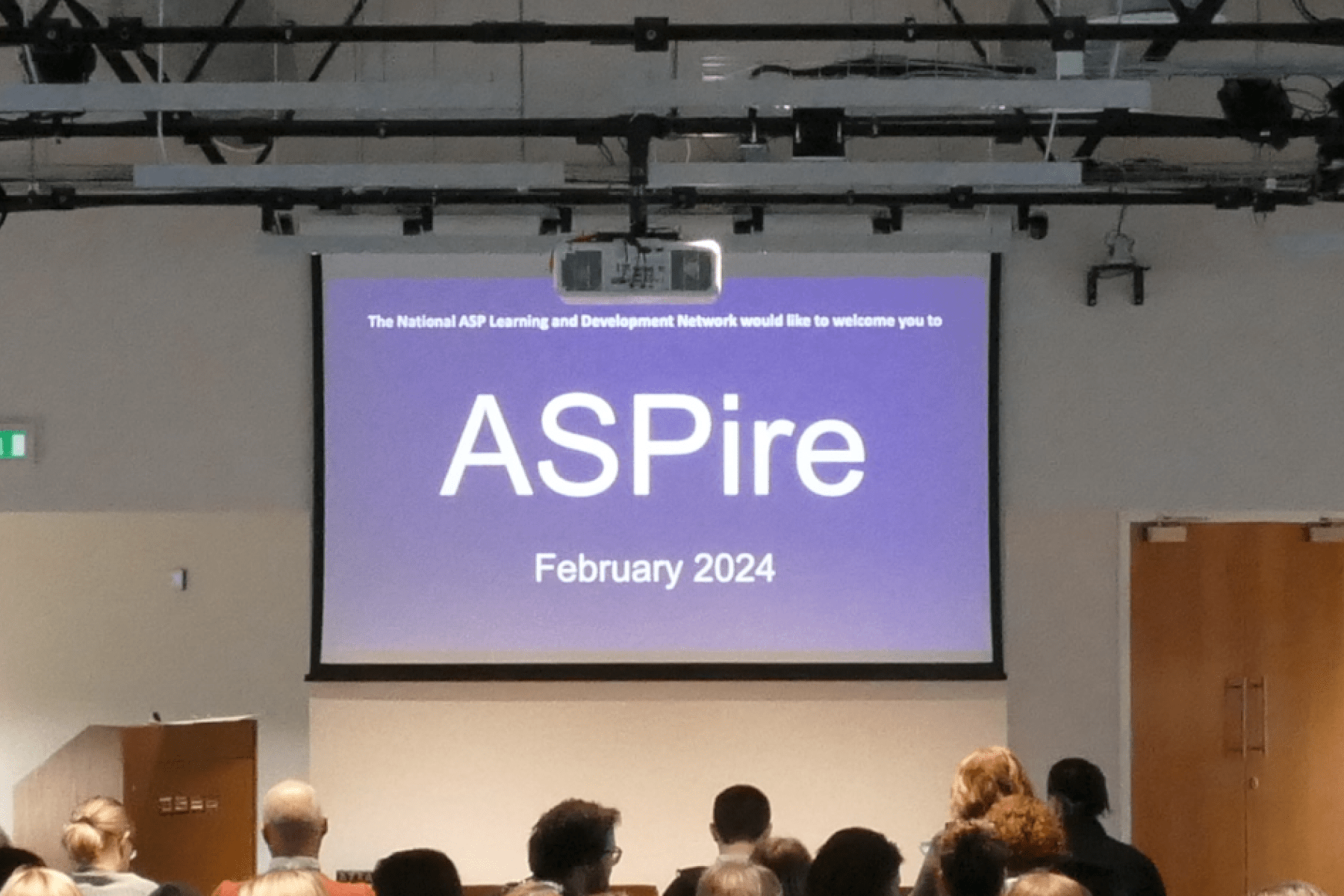 A projector screen with an image of a purple background with the white text ASPire