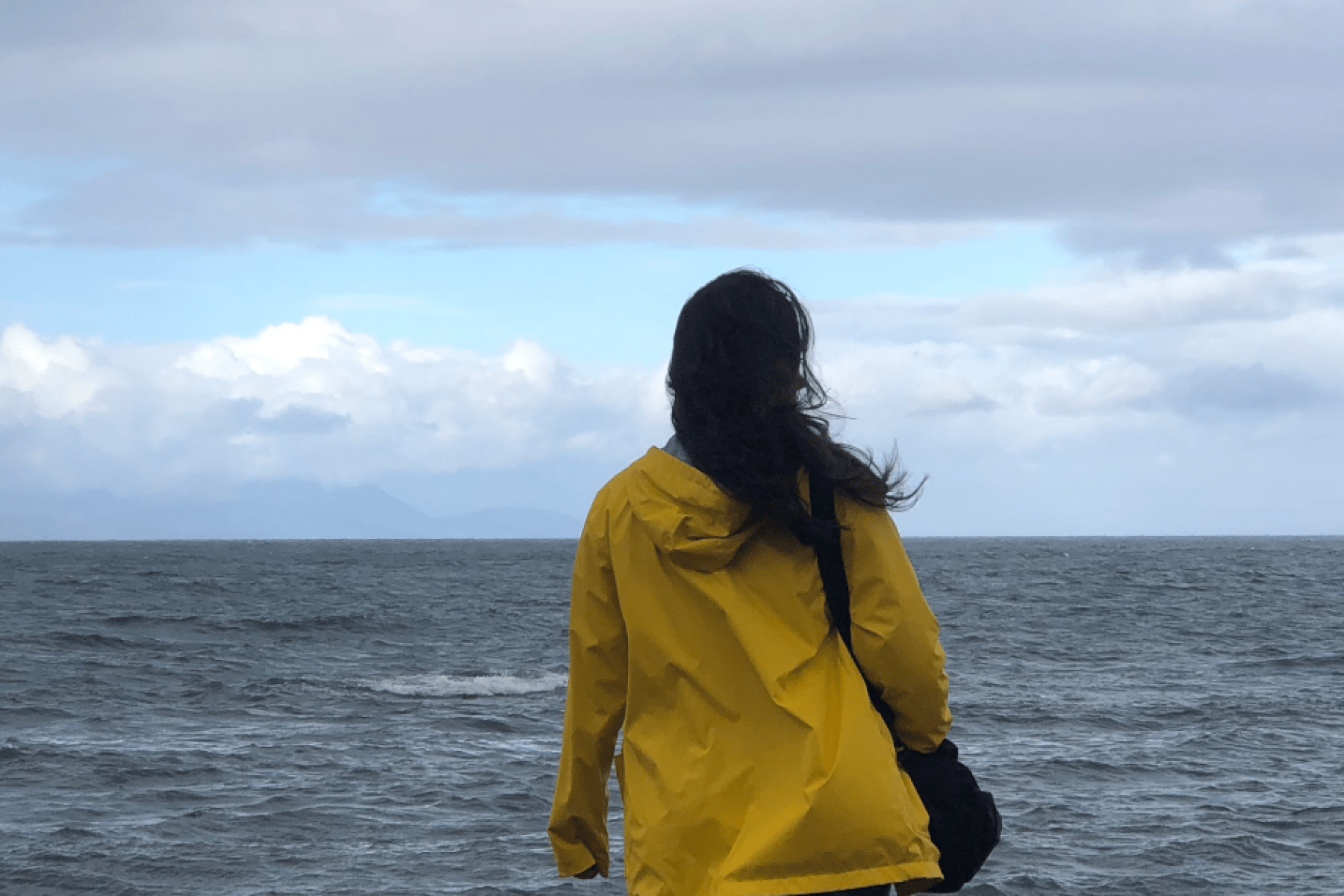 a lady in a yellow raincoat with hair blowing in the wind looking out to sea