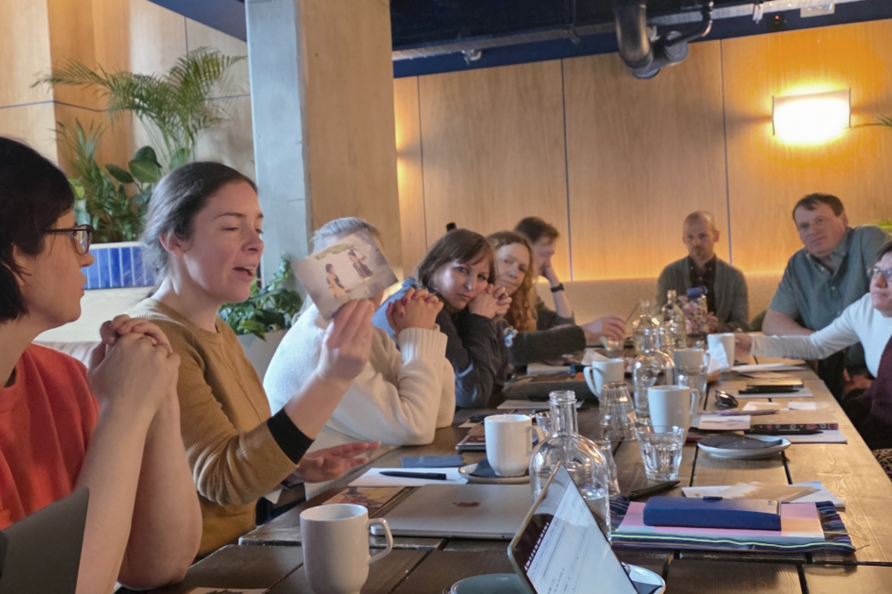 a group of people in discussion around a long table with one person holding a photo