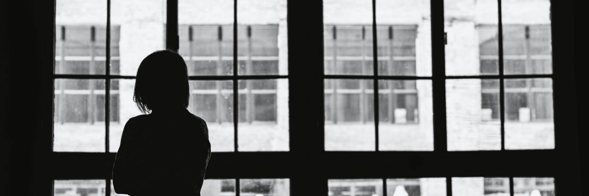 Black and white image of a woman staring out of an industrial looking window