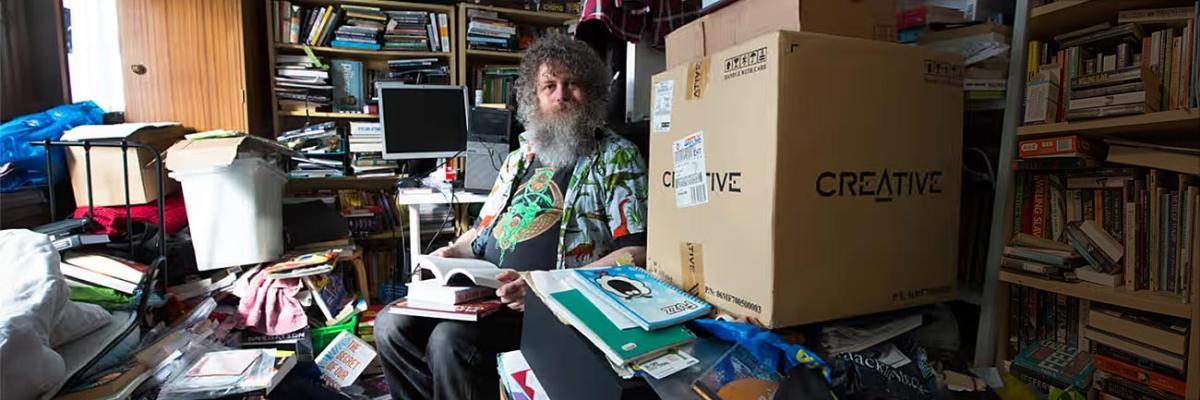 Man seated in bedroom fill with disordered clutter