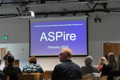 An audience looking at a screen with the word ASPire projected 