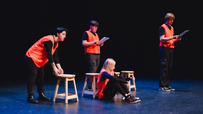 4 people in voluminous ornage vests over all black clothing reading from scripts on a brightly lit sound stage