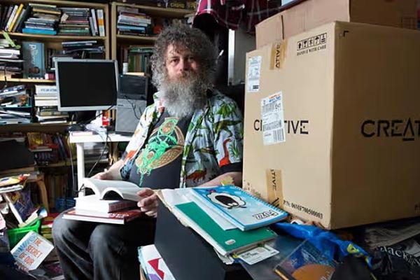Man seated in bedroom fill with disordered clutter